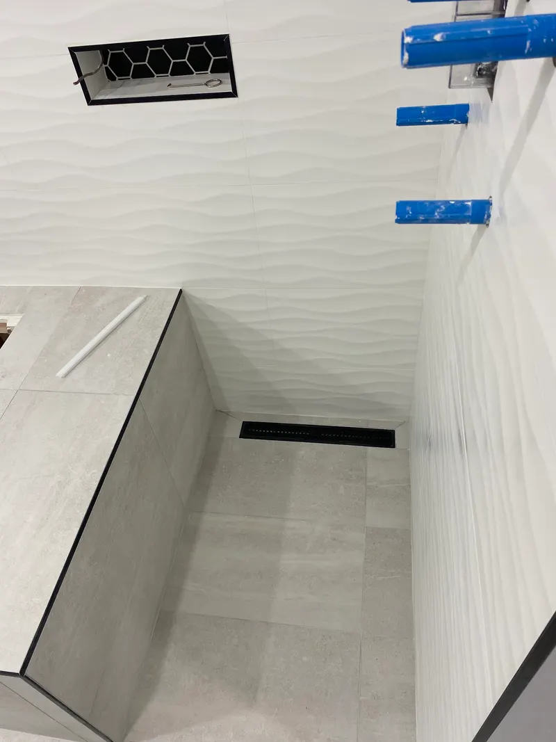 Bathroom renovation by PS Tiling, photo from a Google review: grey porcelain shower floor with a long black drain, white wave-texture walls and a hexagon mosaic niche, mid-renovation.
