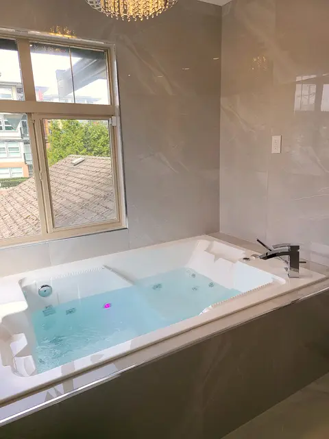Master bathroom renovation in Burquitlam, Coquitlam by PS Tiling: jacuzzi tub filled, with the window view and marble-look porcelain walls, finished.